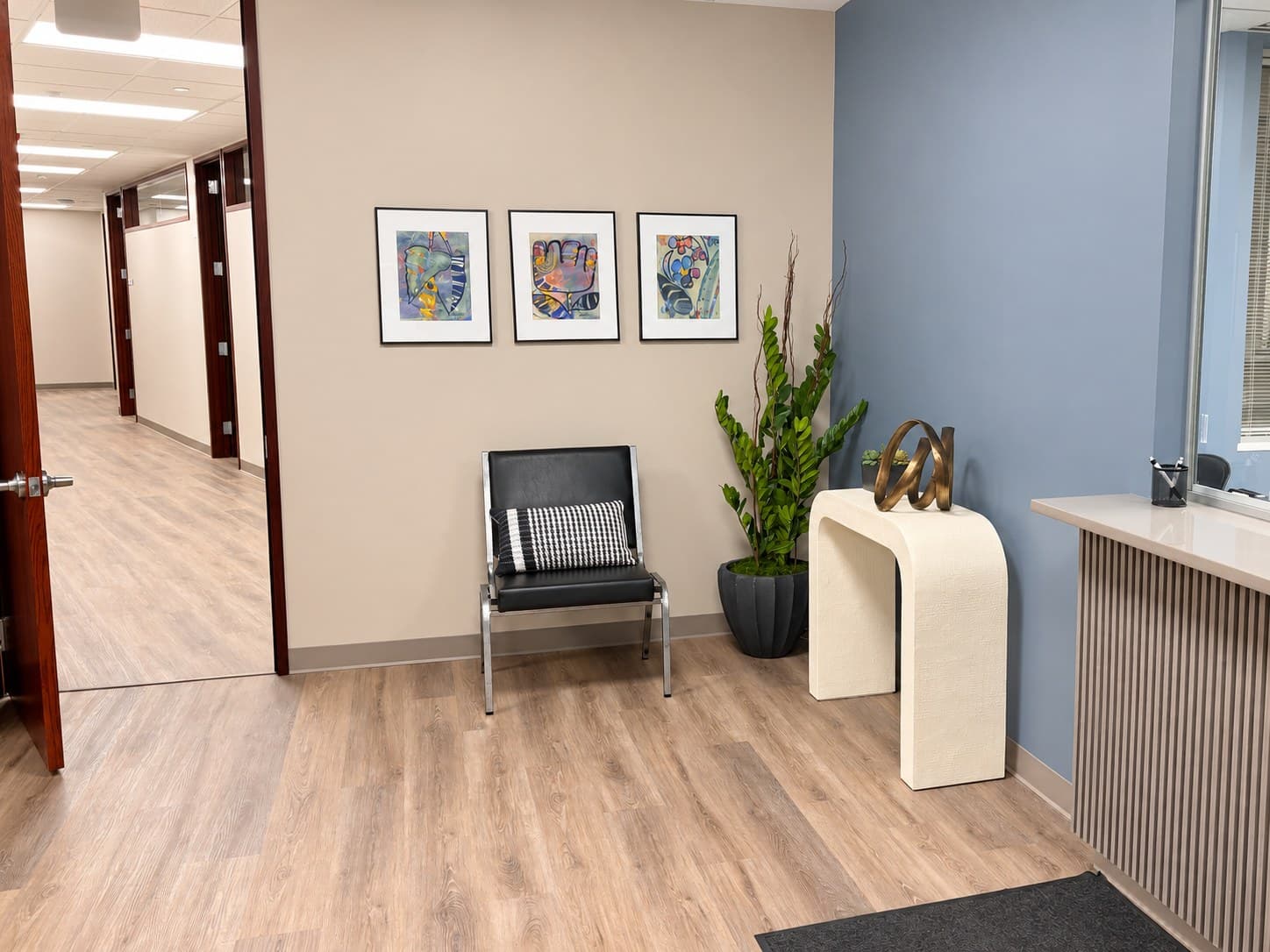 Reception area with a chair, plants, framed artwork, and a glass reception window.